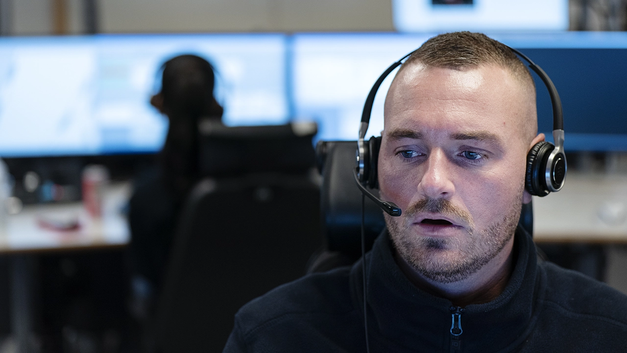 Person med headset sitter i ett larm- eller ledningscenter, med stora skärmar och arbetsstationer synliga i bakgrunden.