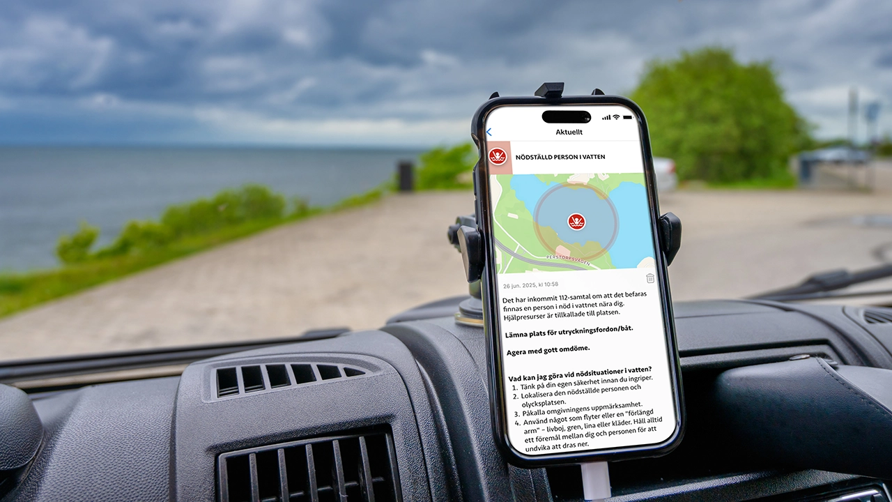 En mobiltelefon sitter fast i en bilhållare på instrumentbrädan och visar 112-appens karta med en markering för ”Nödställd person i vatten”.