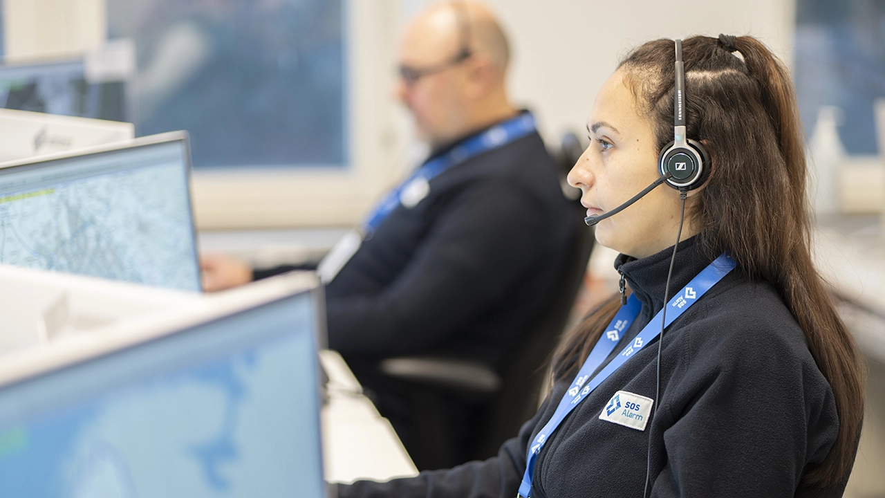 Två personer arbetar vid datorer i en larmcentral, båda bär headset och kläder med SOS Alarms logotyp.
