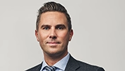 Person iförd mörk kavaj, ljus skjorta och randig slips står framför en ljus, neutral bakgrund.