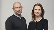 Två personer står bredvid varandra mot en neutral bakgrund, båda klädda i mörka överdelar med ljusare skjortor under.