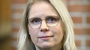 Person med ljust axellångt hår bär en grön tröja och en prästkrage, stående inomhus framför en murad vägg.
