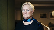 Person i mörk överdel med blåmönstrad scarf står inomhus mot en mjukt belyst bakgrund.