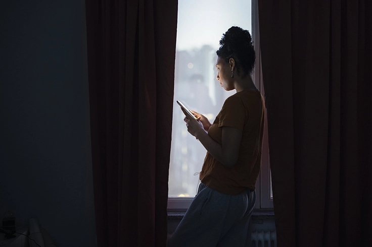 Person står vid ett fönster i ett mörkt rum och håller en mobiltelefon som lyser upp siluetten mot det svaga ljuset utifrån.