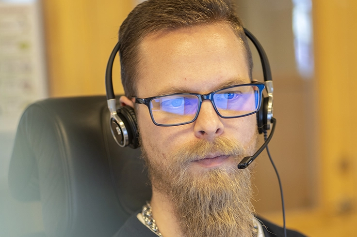Person med headset sitter i en stol, med kortklippt hår och skägg synligt, i en miljö med varmt ljus.