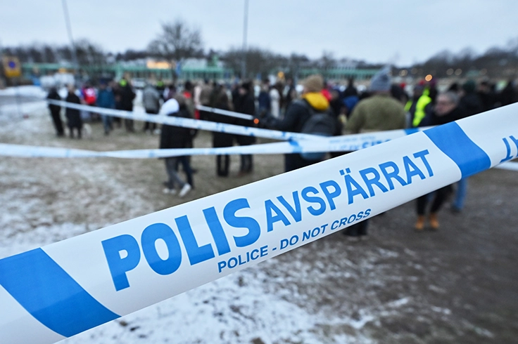 Polisavspärrning med blåvit tejp i förgrunden och en grupp människor som står samlade på en snötäckt plats i bakgrunden.