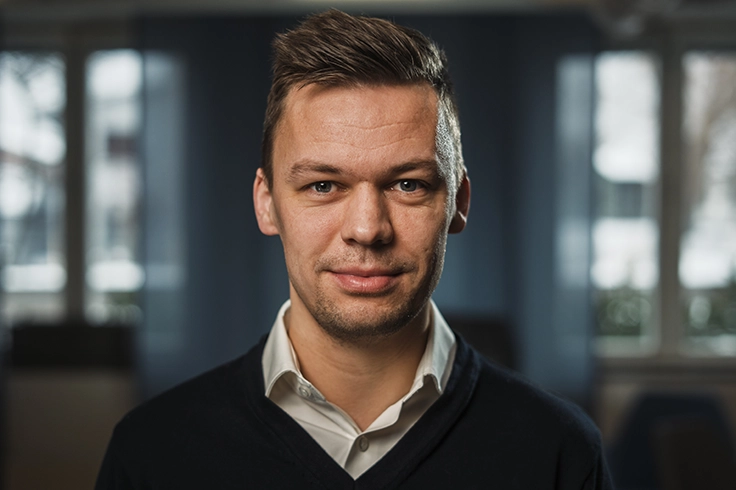 Person klädd i mörk tröja och ljus skjorta står inomhus framför en bakgrund med stora fönster och dämpad belysning.Ge feedback om BizChat