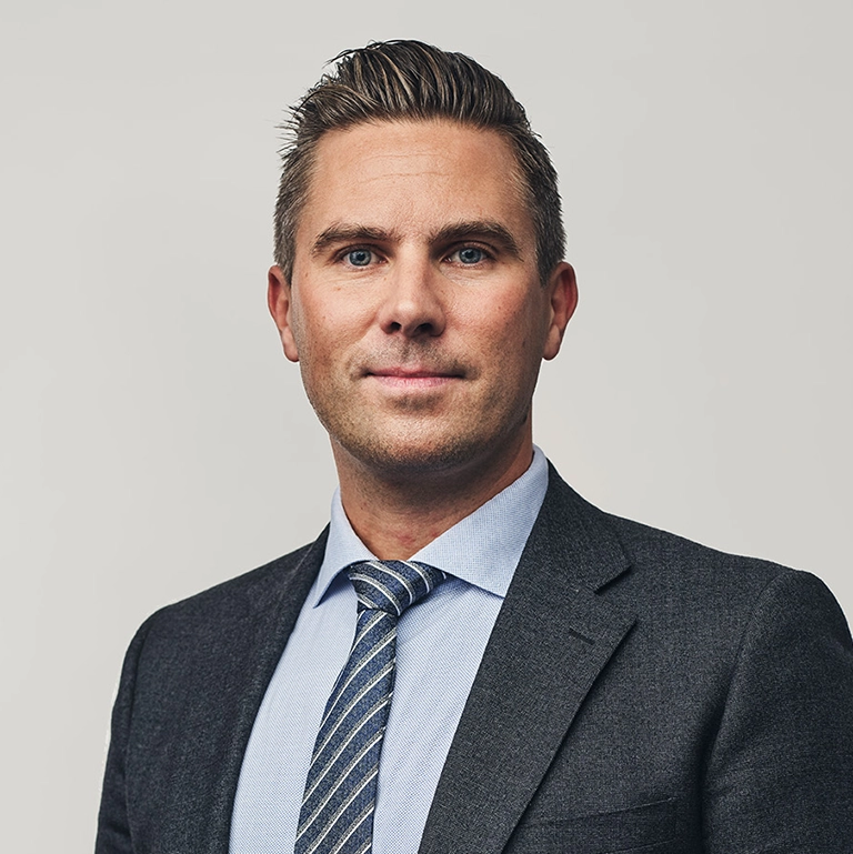 Person iförd mörk kavaj, ljus skjorta och randig slips står framför en ljus, neutral bakgrund.
