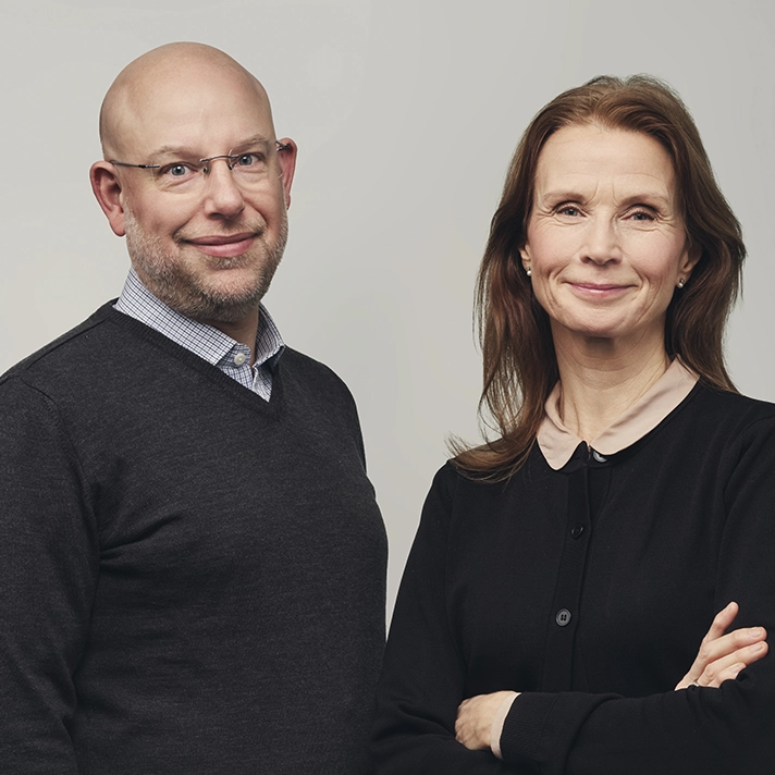 Två personer står bredvid varandra mot en neutral bakgrund, båda klädda i mörka överdelar med ljusare skjortor under.