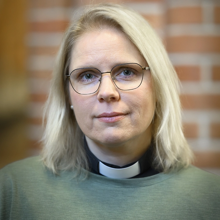 Person med ljust axellångt hår bär en grön tröja och en prästkrage, stående inomhus framför en murad vägg.
