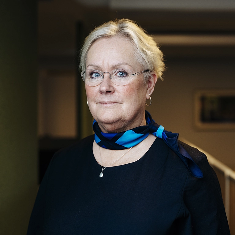 Person i mörk överdel med blåmönstrad scarf står inomhus mot en mjukt belyst bakgrund.